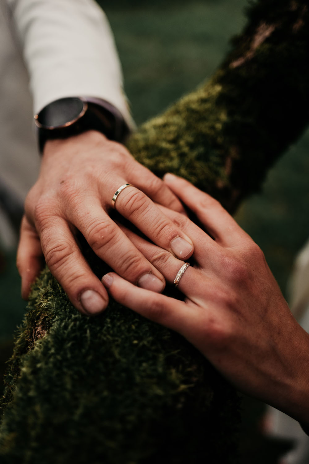 Mains d’un couple marié portant leurs alliances posées sur un tronc recouvert de mousse.