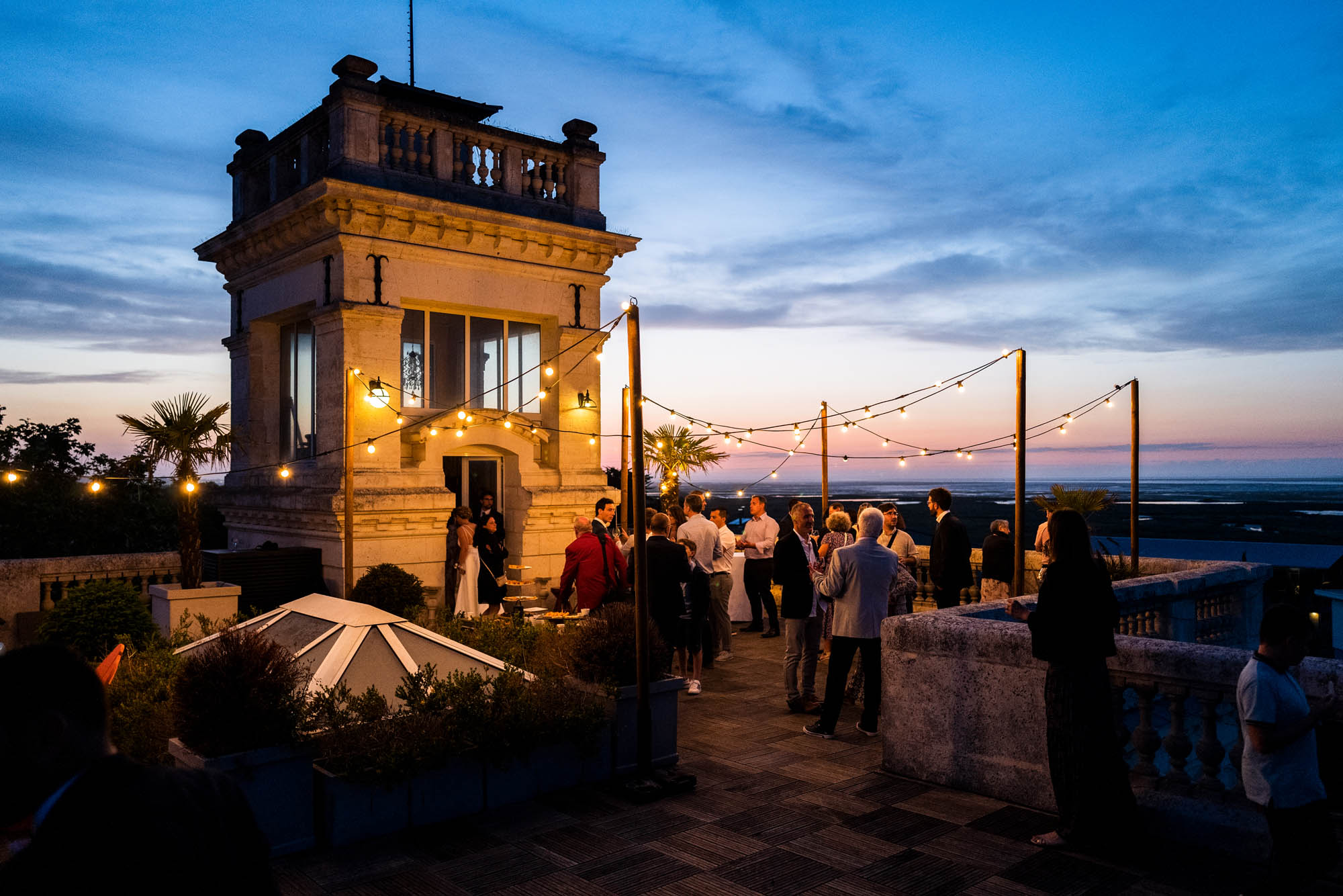 Réception de mariage sur une terrasse de château au coucher du soleil, invités discutant sous des guirlandes lumineuses.