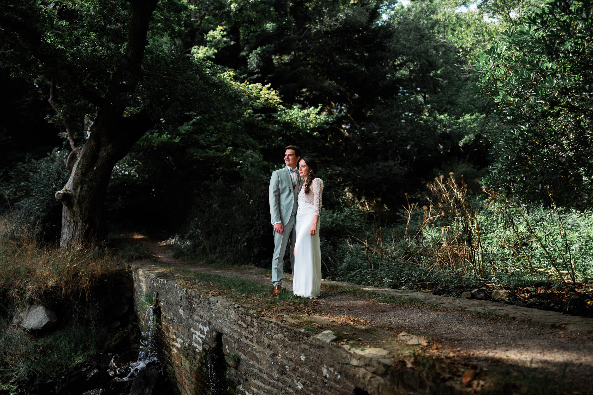 Couple de mariés debout sur un petit pont en pierre au cœur d’une forêt verdoyante bretonne.