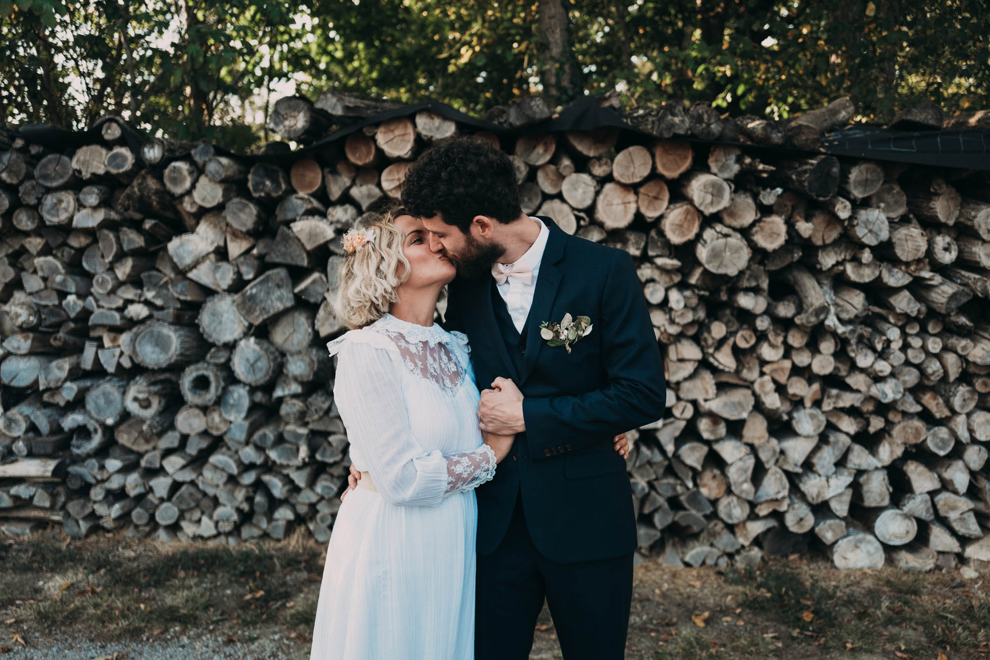 Couple de mariés élégant posant devant un mur de bûches dans un cadre champêtre et naturel.