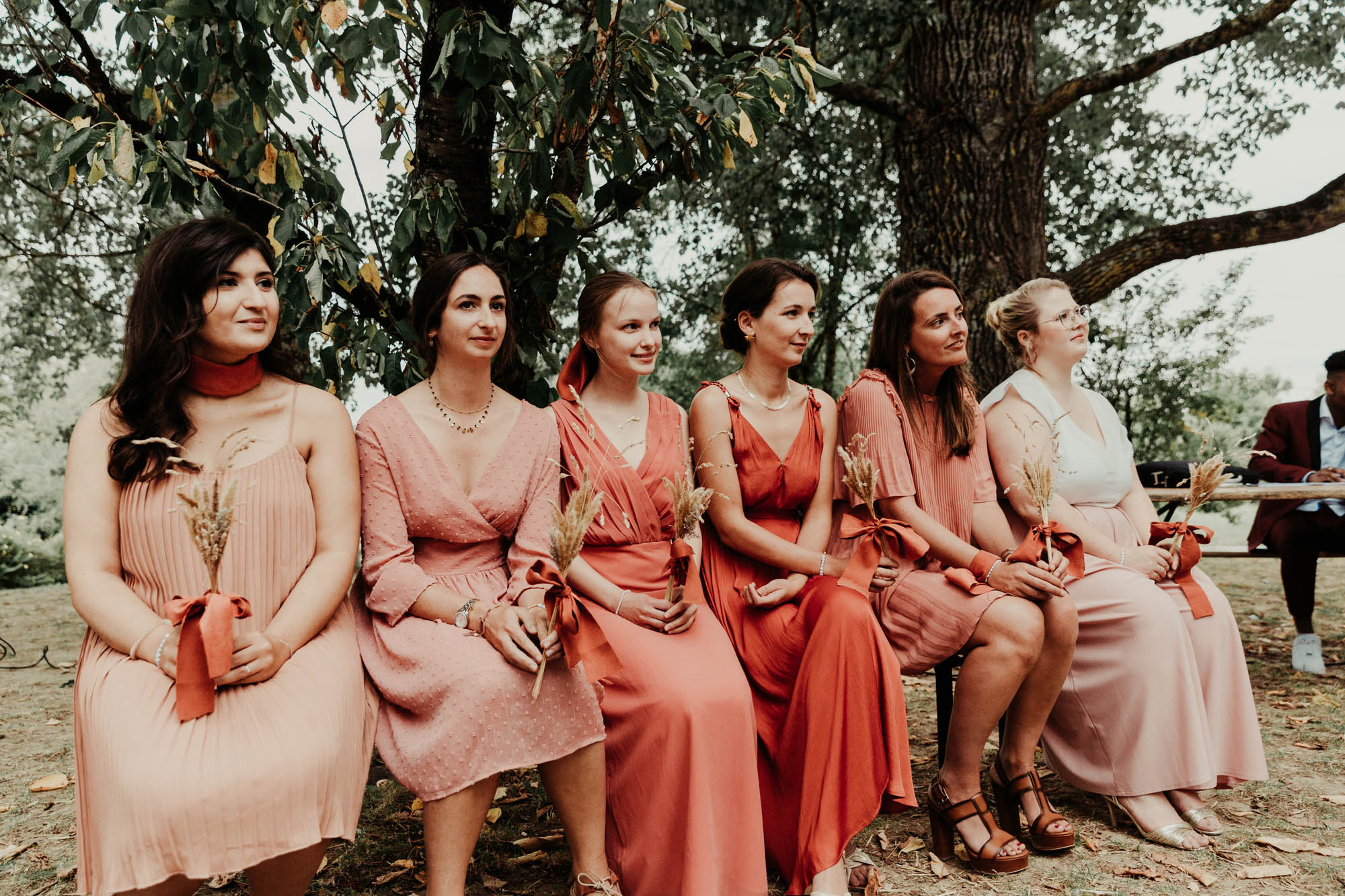 Demoiselles d’honneur assises en extérieur lors d’un mariage, portant des robes roses assorties et tenant des bouquets de fleurs séchées.