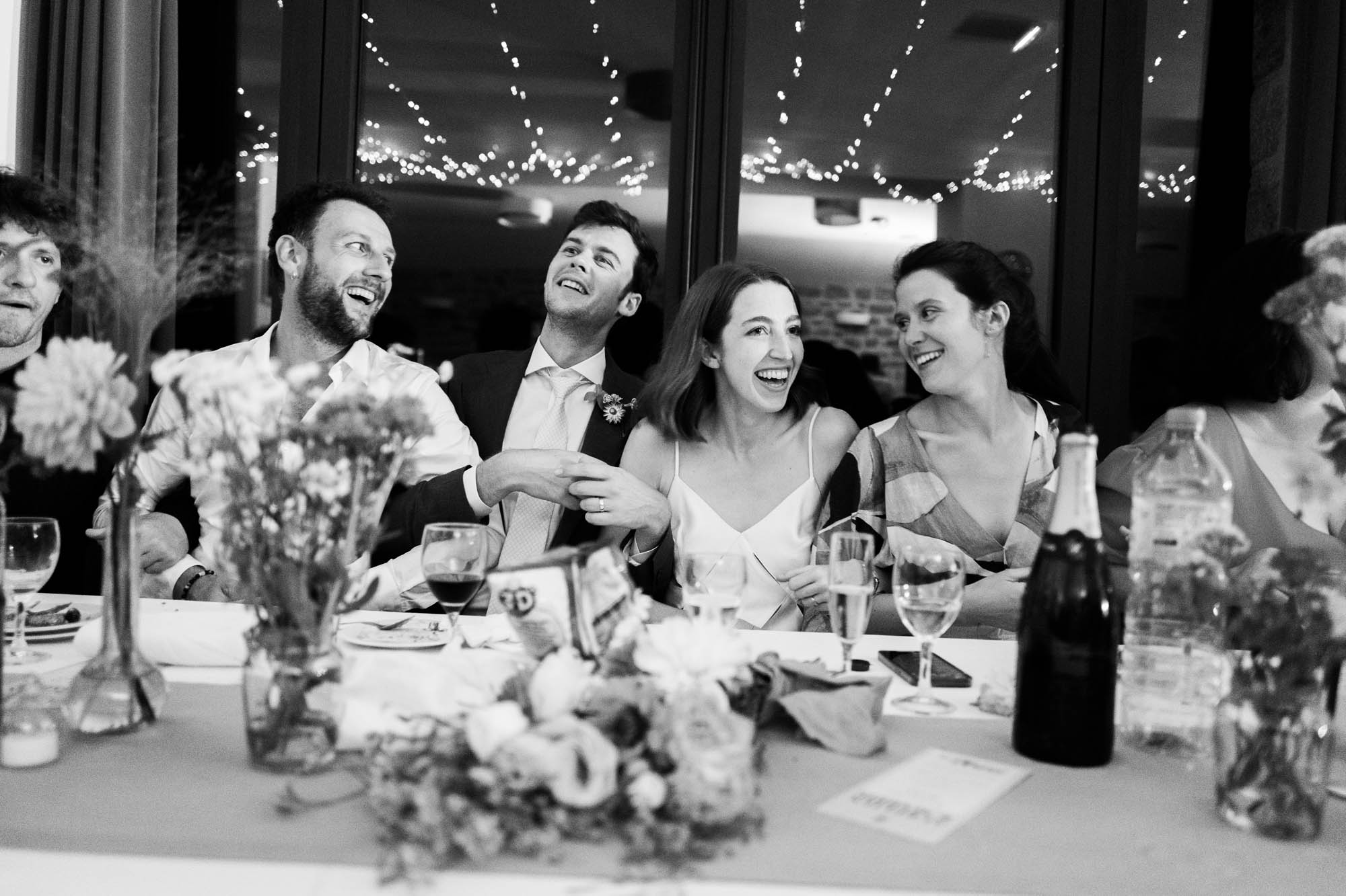 Groupe de convives assis à une table élégamment décorée lors d’un dîner de mariage en noir et blanc, avec verres, bouteilles et compositions florales.