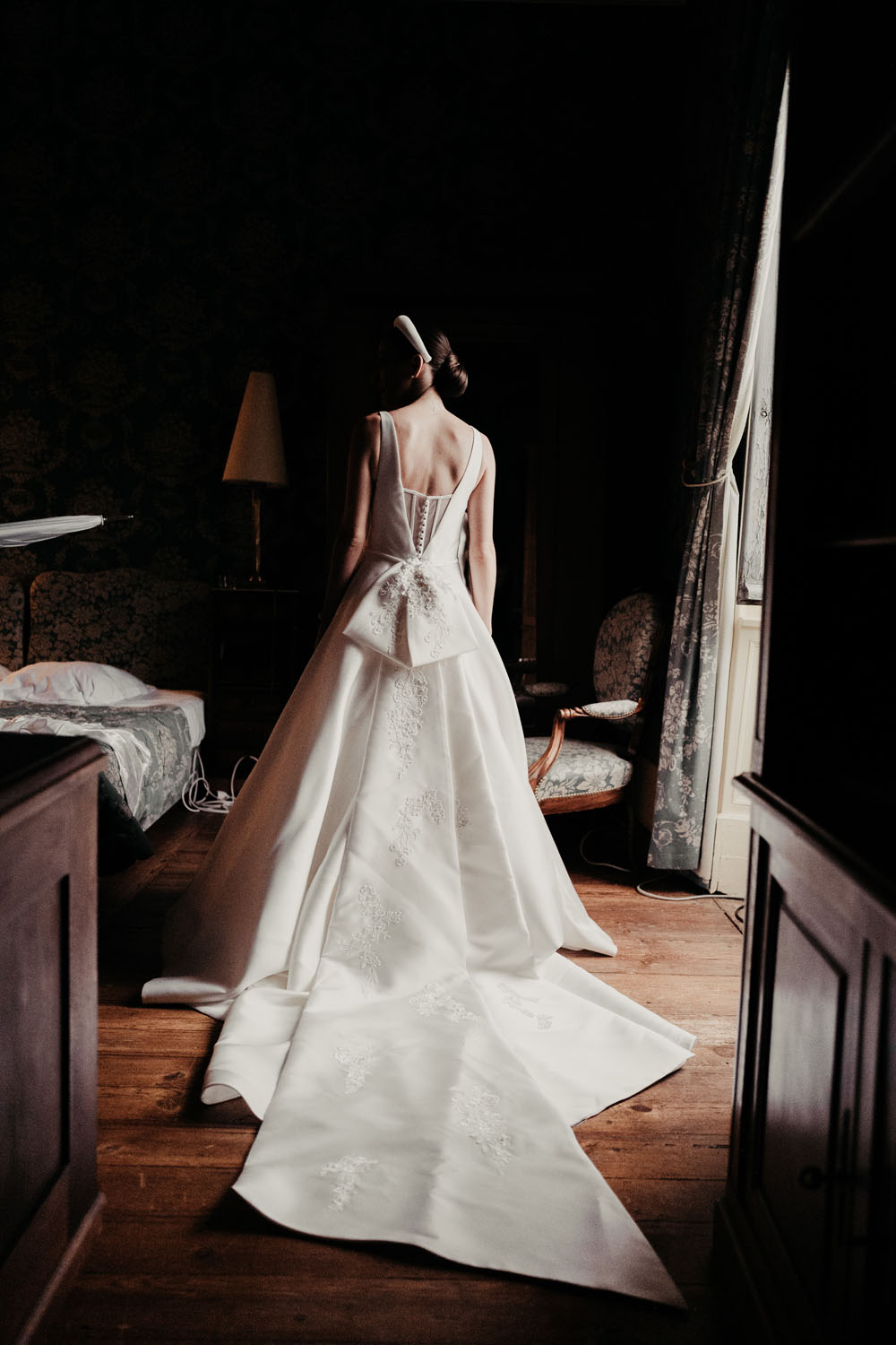 Vue de dos d’une mariée portant une robe blanche à longue traîne dans une chambre lors des préparatifs de mariage.