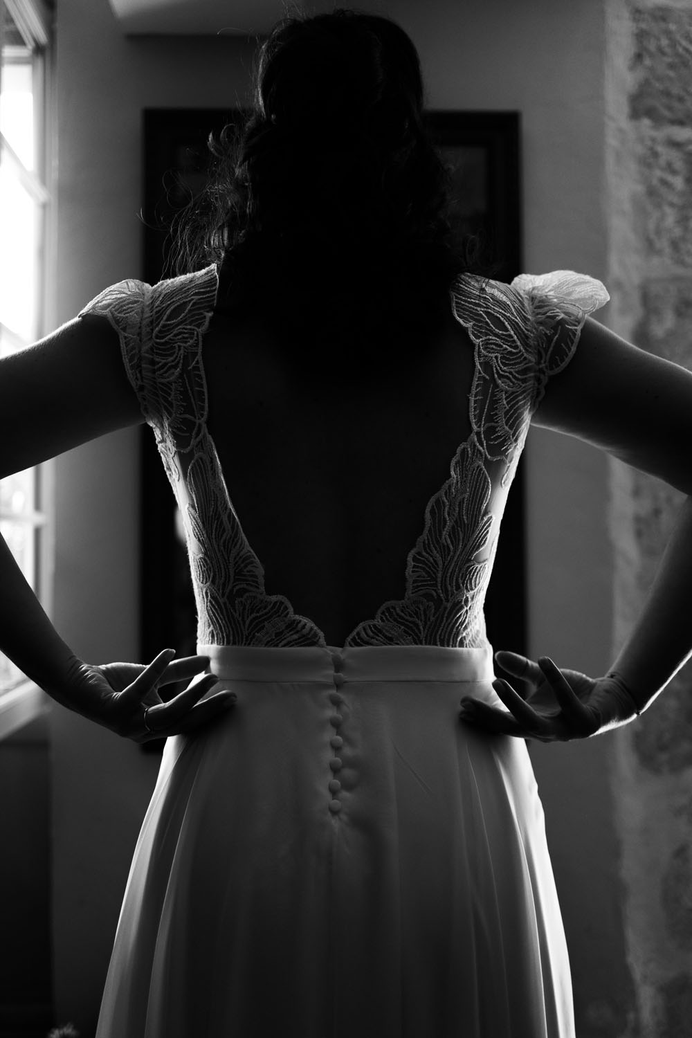 Vue de dos d’une mariée en robe de dentelle élégante, les mains posées sur les hanches, éclairée par la lumière d’une fenêtre.