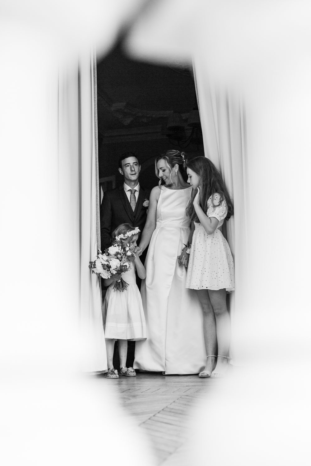 Groupe familial lors d’un mariage, composé d’un couple en tenue de cérémonie avec deux enfants, debout à l’entrée d’une pièce, bouquet de fleurs à la main, photo en noir et blanc.