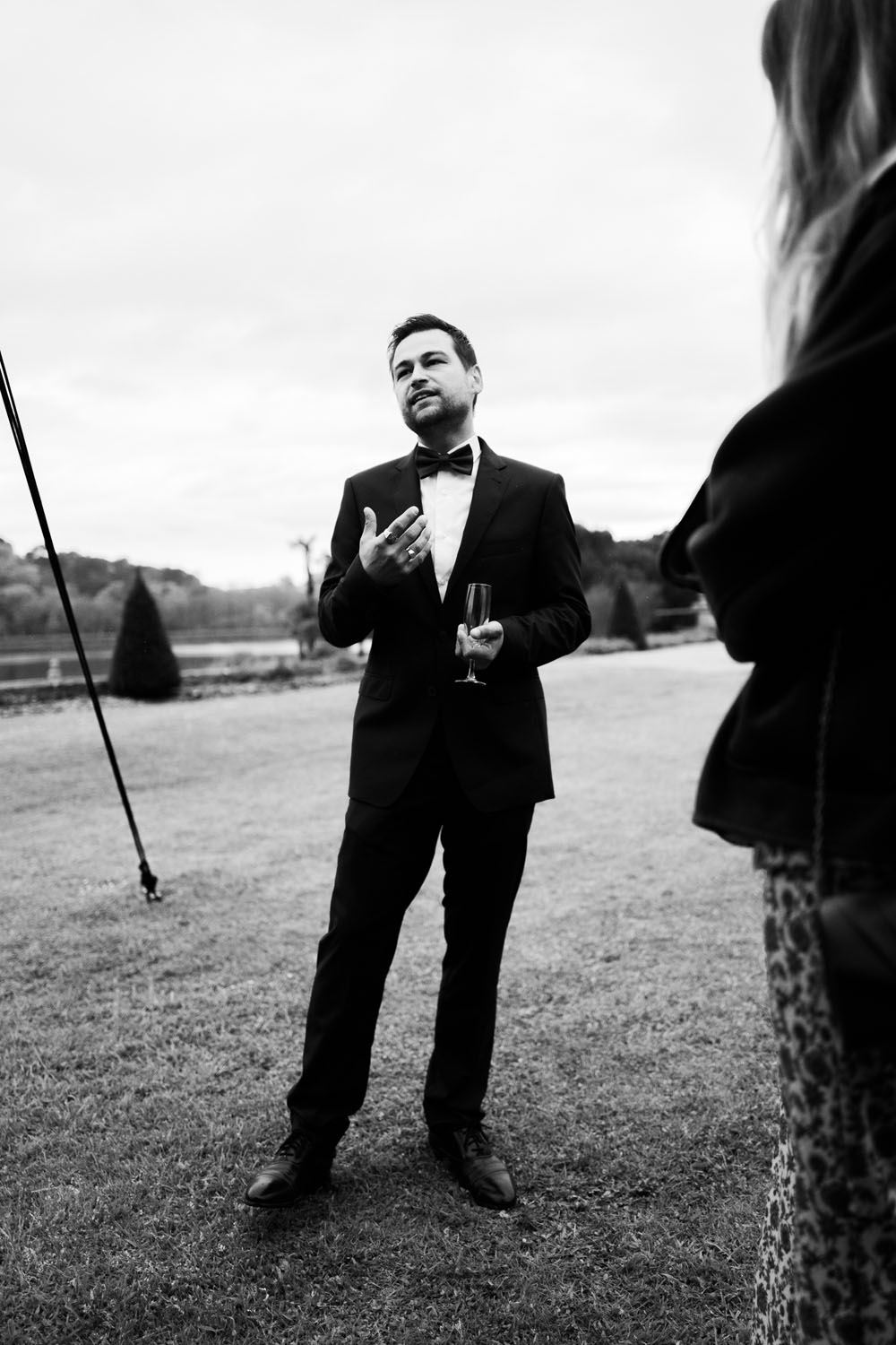 Homme en costume avec nœud papillon tenant un verre de champagne lors d’une réception en extérieur.