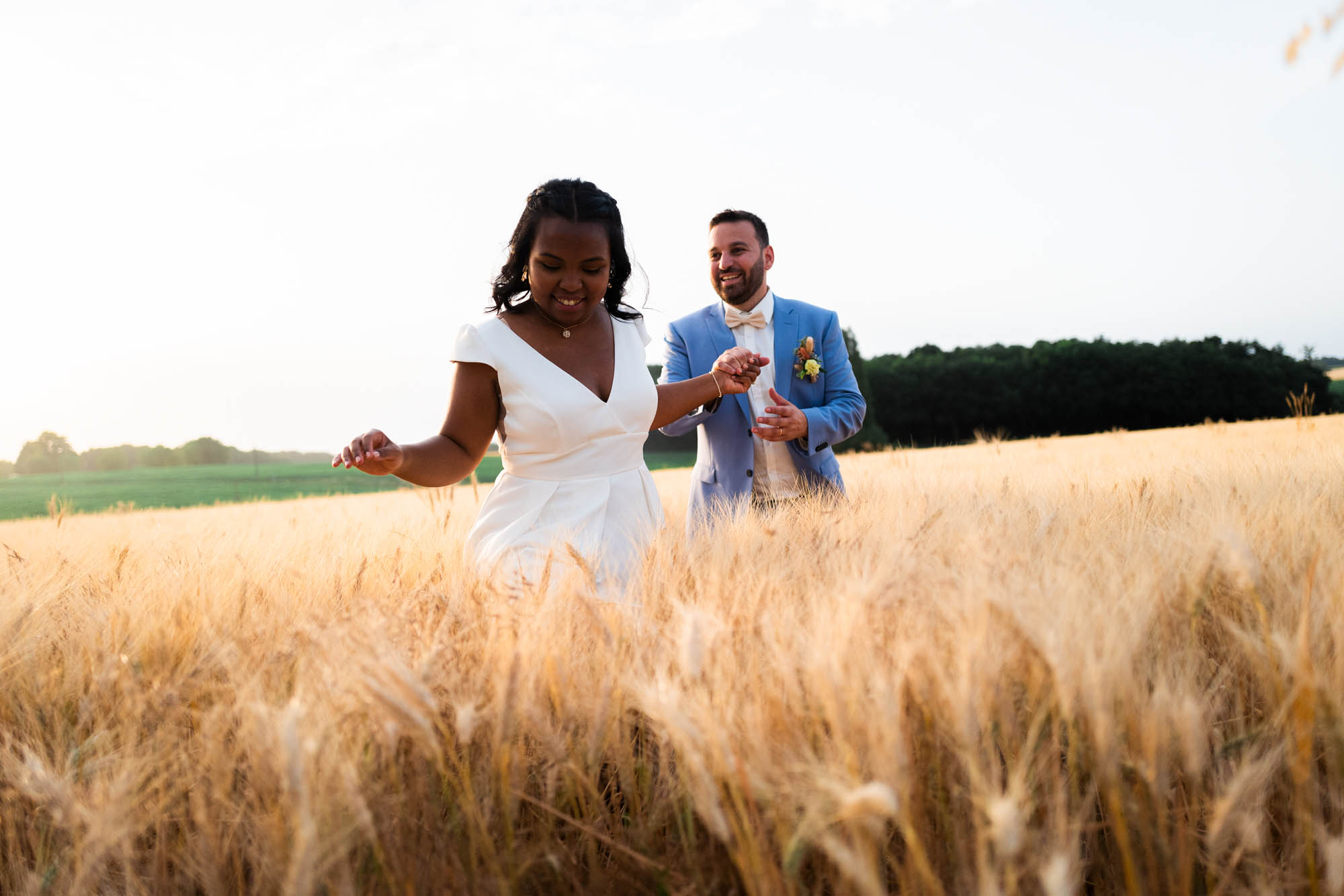 Couple de mariés marchant dans un champ de blé au coucher du soleil, ambiance naturelle et estivale.