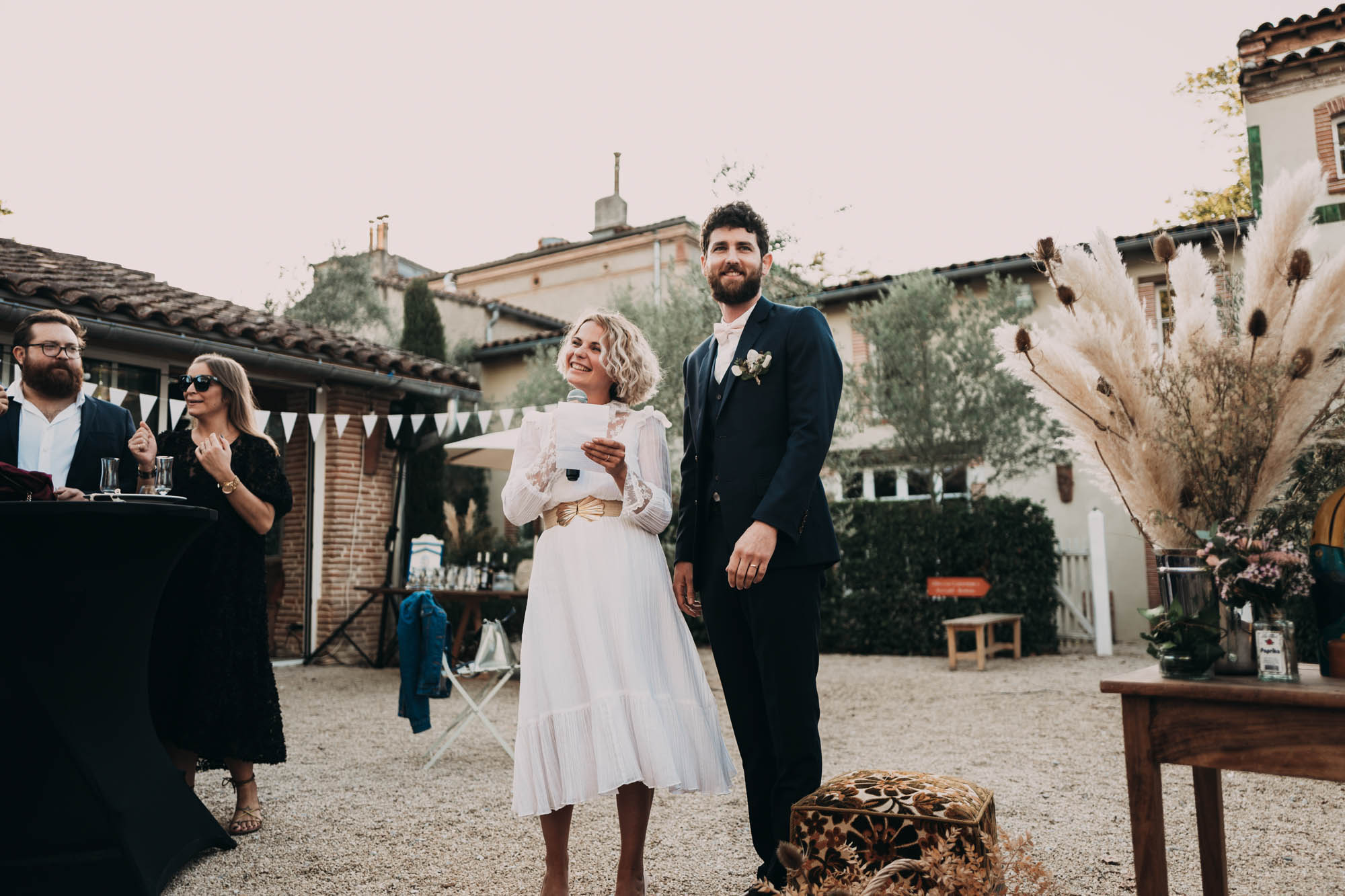 Un couple habillé pour un mariage dans une cour extérieure décorée de pampas avec des invités, ambiance champêtre et bohème