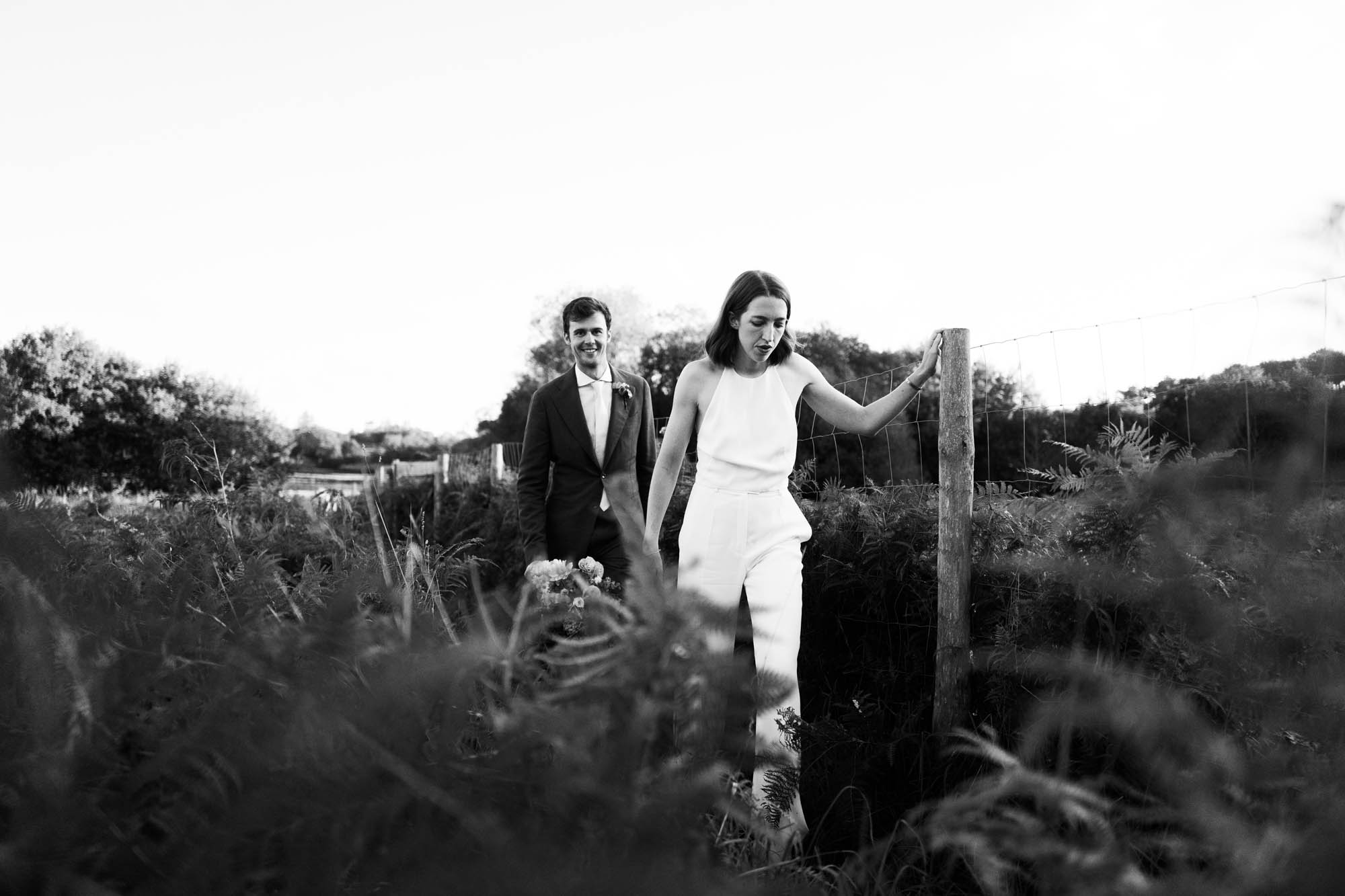 Couple de mariés marchant côte à côte dans un champ en noir et blanc, la mariée tenant un bouquet de fleurs.