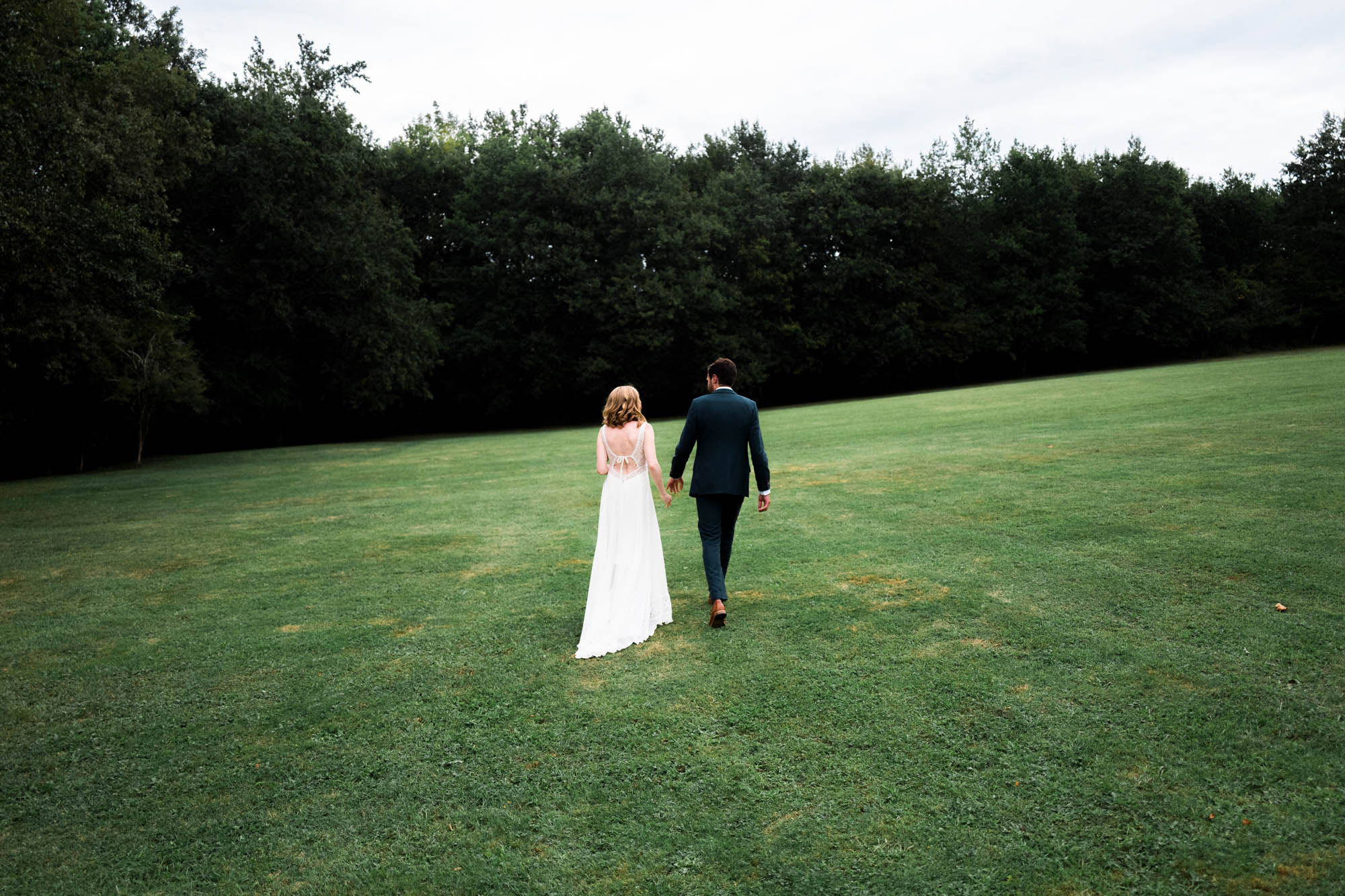 Mariés de dos marchant main dans la main sur un grand champ vert entouré d’arbres.