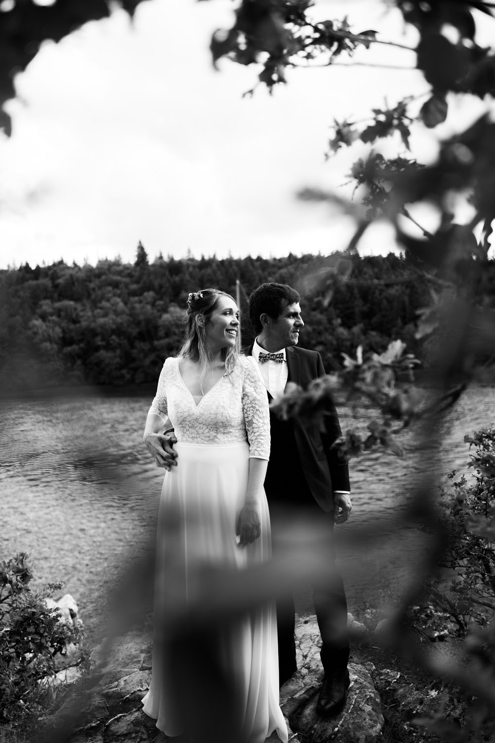 Couple de mariés debout au bord d’une rivière, entourés de végétation, capturé en noir et blanc.