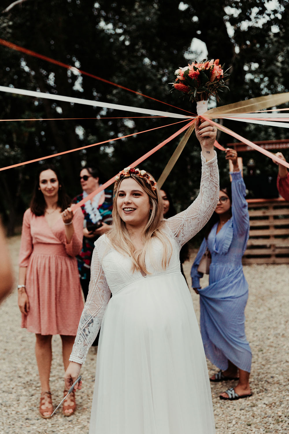 Mariée en robe blanche tenant un bouquet de fleurs lors du jeu des rubans au mariage en plein air.