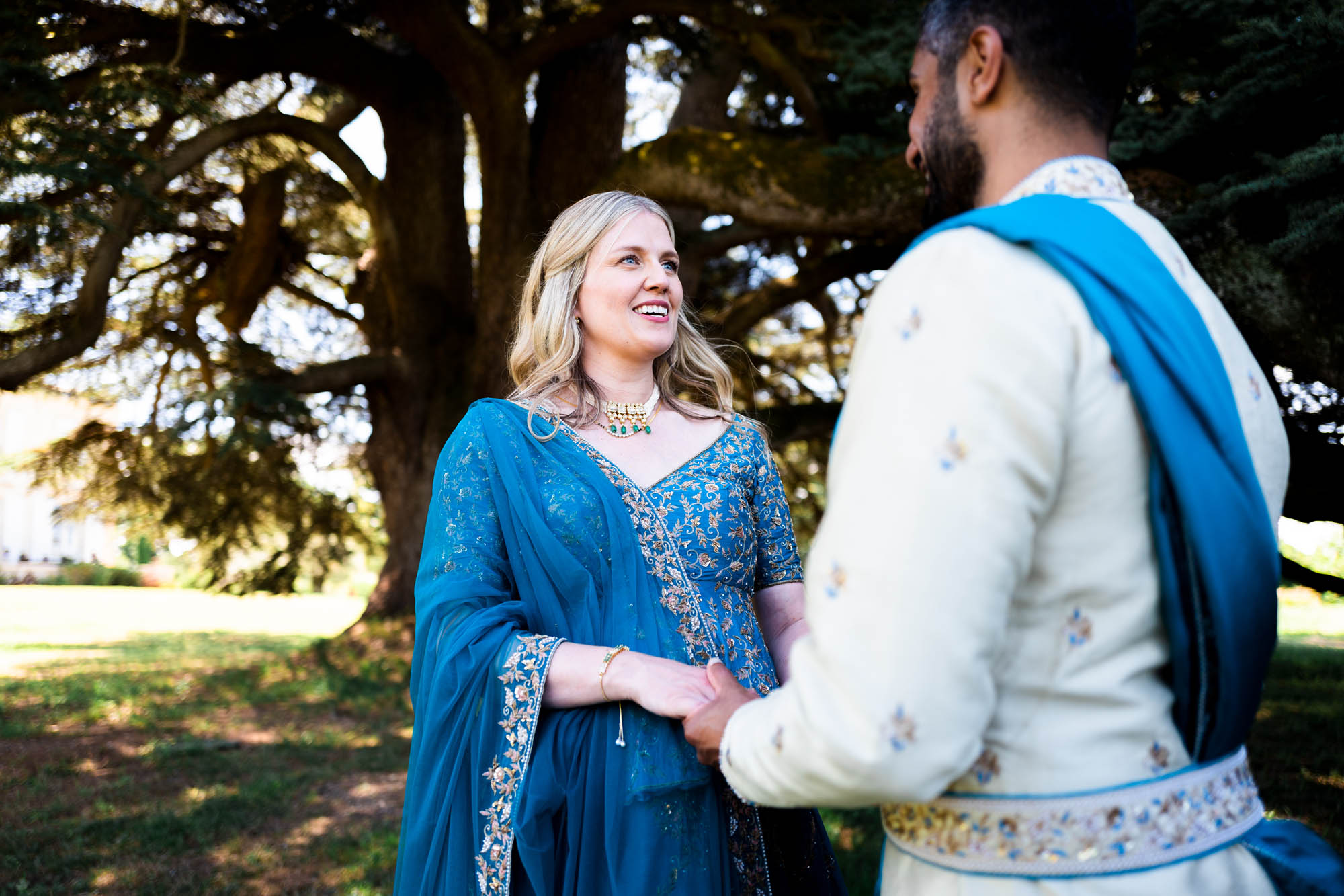 Couple en tenues traditionnelles indiennes tenant leurs mains, debout sous un grand arbre en plein air.