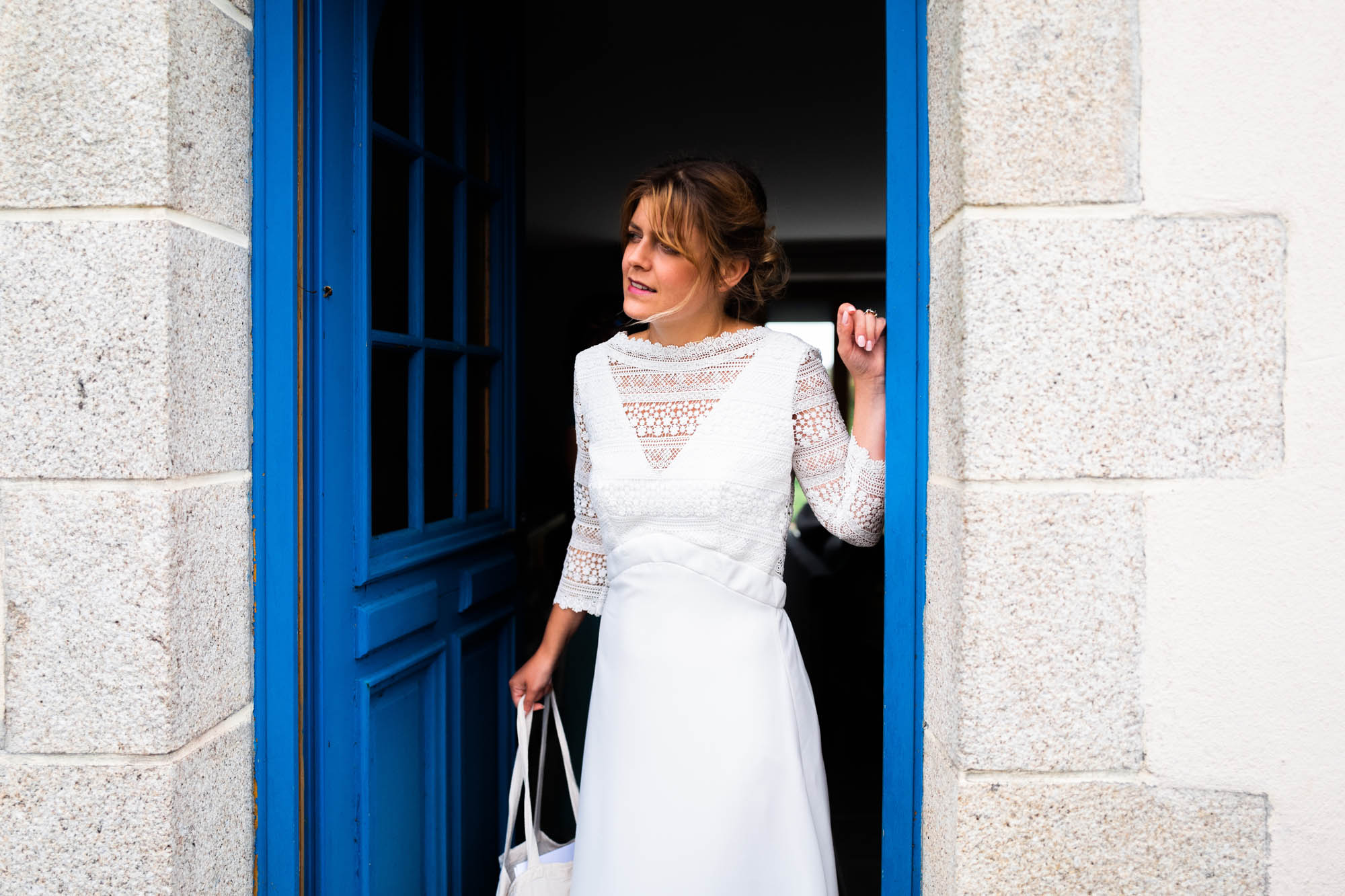 Mariée en robe blanche entrant ou sortant par une porte bleue encadrée de pierres bretonnes.