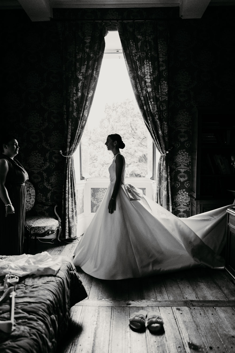 Mariée debout en robe blanche devant une fenêtre, avec une ambiance intime et élégante dans une chambre.