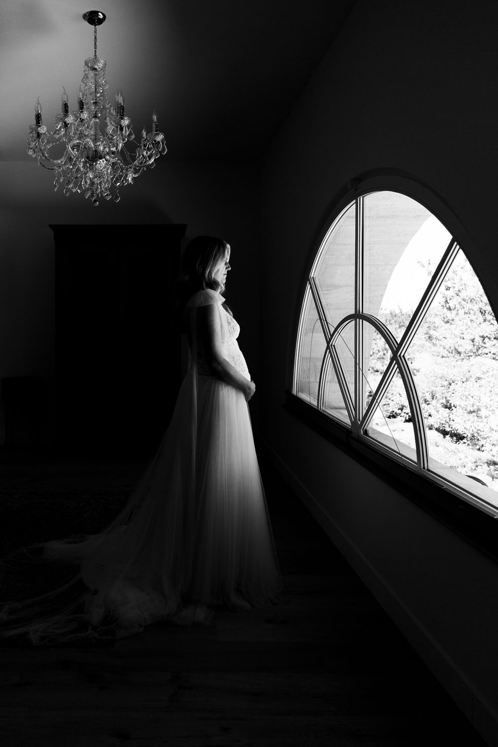 Mariée en robe blanche debout près d’une grande fenêtre arquée, regardant à l’extérieur, éclairée par la lumière naturelle.