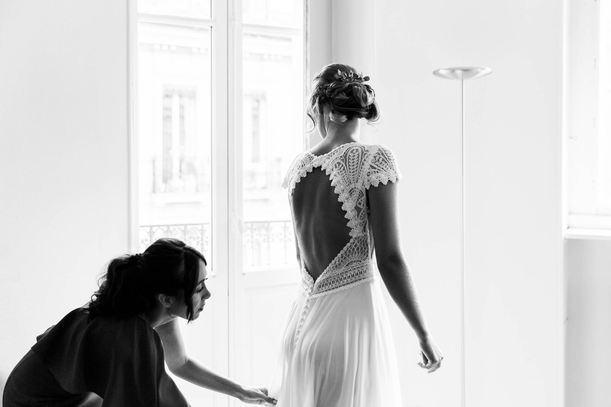Mariée en robe de dentelle dos nu se faisant aider pour boutonner sa robe devant une fenêtre.