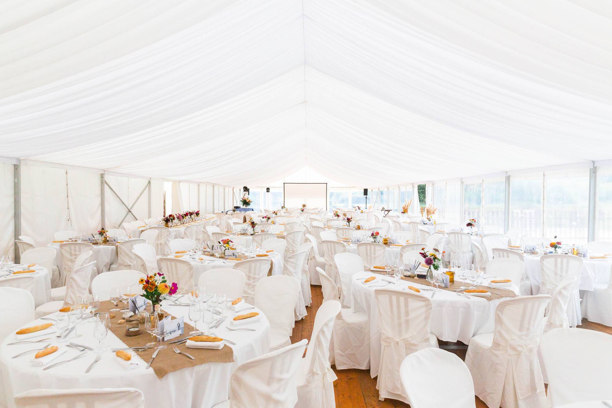 Salle de réception de mariage sous une grande tente blanche, tables rondes décorées de fleurs et de vaisselle élégante.