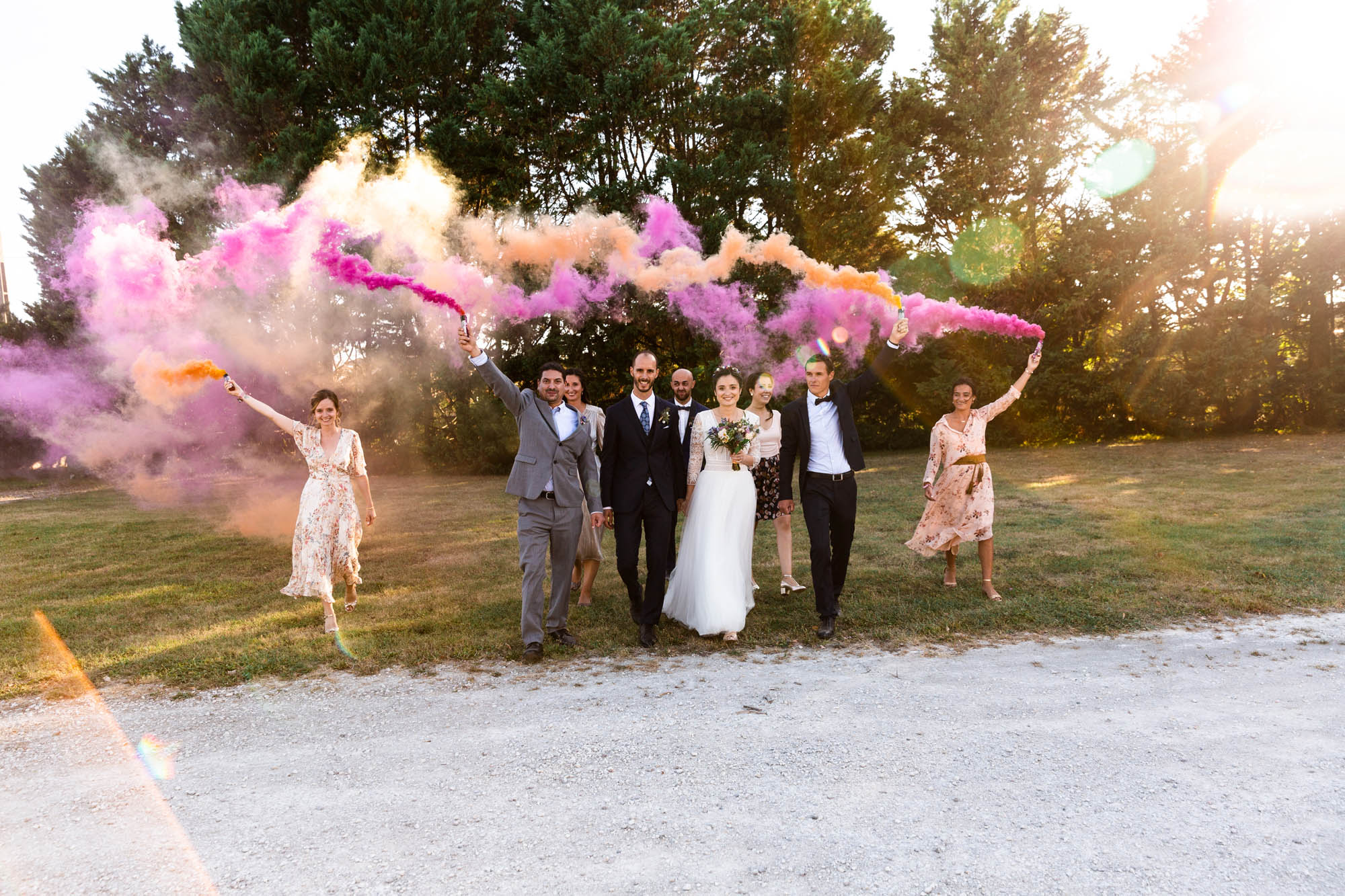 Groupe de mariage avançant ensemble à l’extérieur, tenant des fumigènes colorés rose et orange sous le soleil.