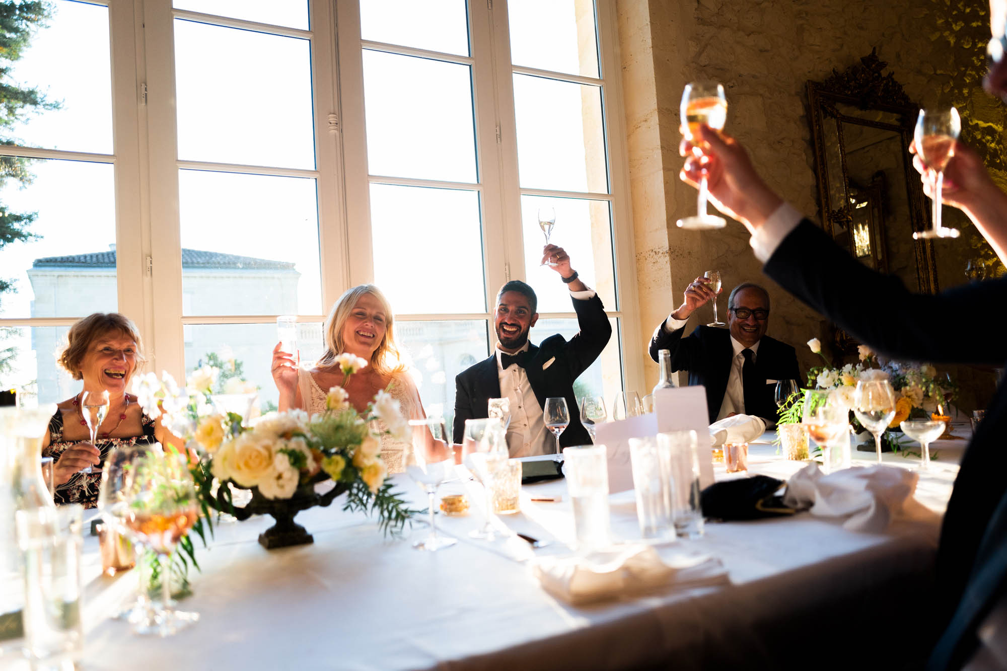 Invités levant leur verre pour un toast lors d’un repas de mariage, table décorée de fleurs, grande fenêtre en arrière-plan.
